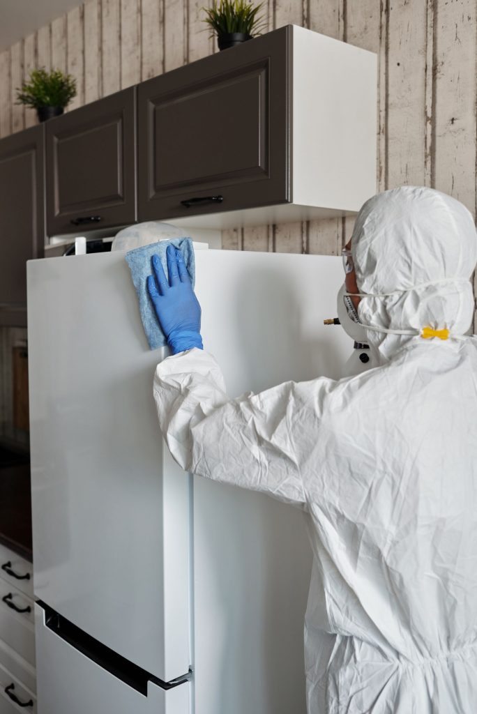 Cleaner in protective clothing disinfecting a white refrigerator