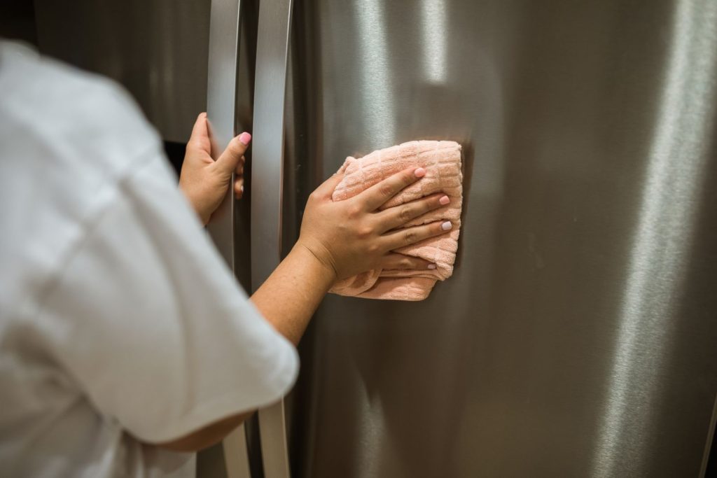 Hands wiping the stainless steel door of a refrigerator with a cloth